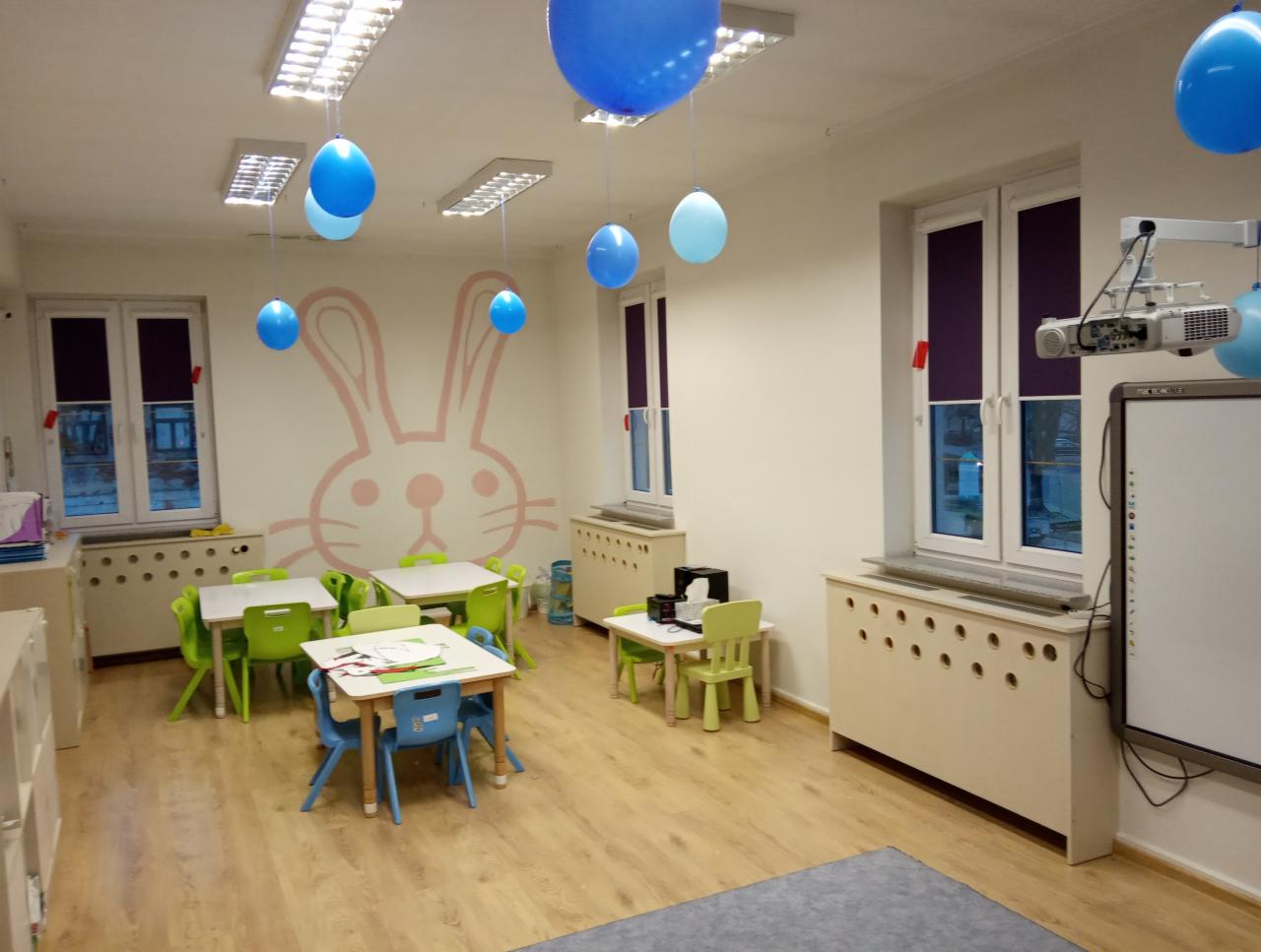 Wnętrze jasnej sali przedszkolnej z kolorowymi krzesłami i stolikami, dekoracjami z niebieskich balonów i rysunkiem królika na ścianie, okna z fioletowymi przesłonami.