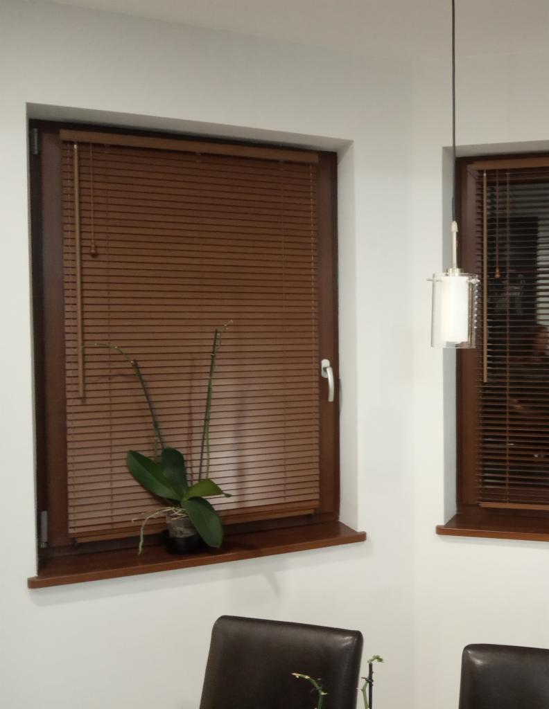 Brązowe żaluzje poziome w drewnianej ramie okiennej, z rośliną doniczkową na parapecie i lampą wiszącą w tle.