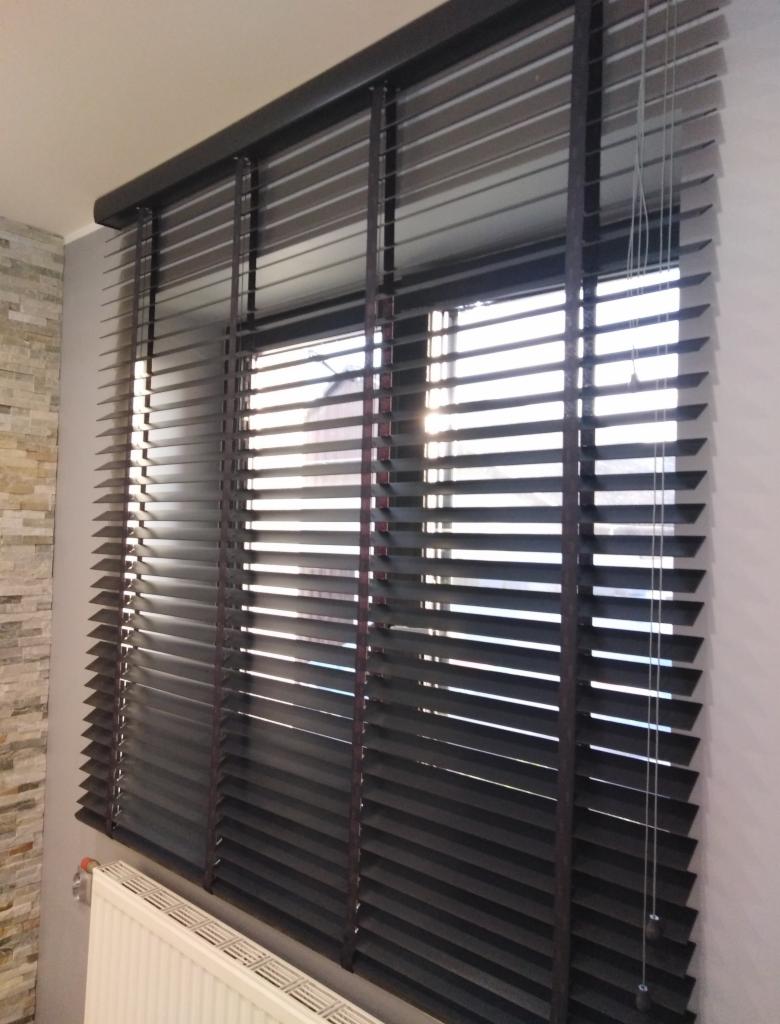Ciemnobrązowe żaluzje poziome z szerokimi lamelami zamontowane w oknie, widoczny fragment grzejnika pod oknem i dekoracyjna ściana z kamienia w tle.