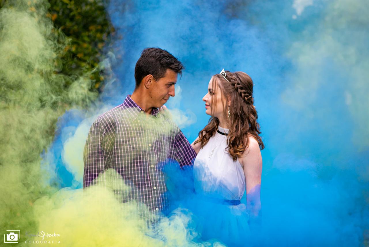 Para spogląda na siebie w obłokach niebieskiego i zielonego dymu; dziewczyna w tiulowej sukience i diademie, chłopak w koszuli w kratę.