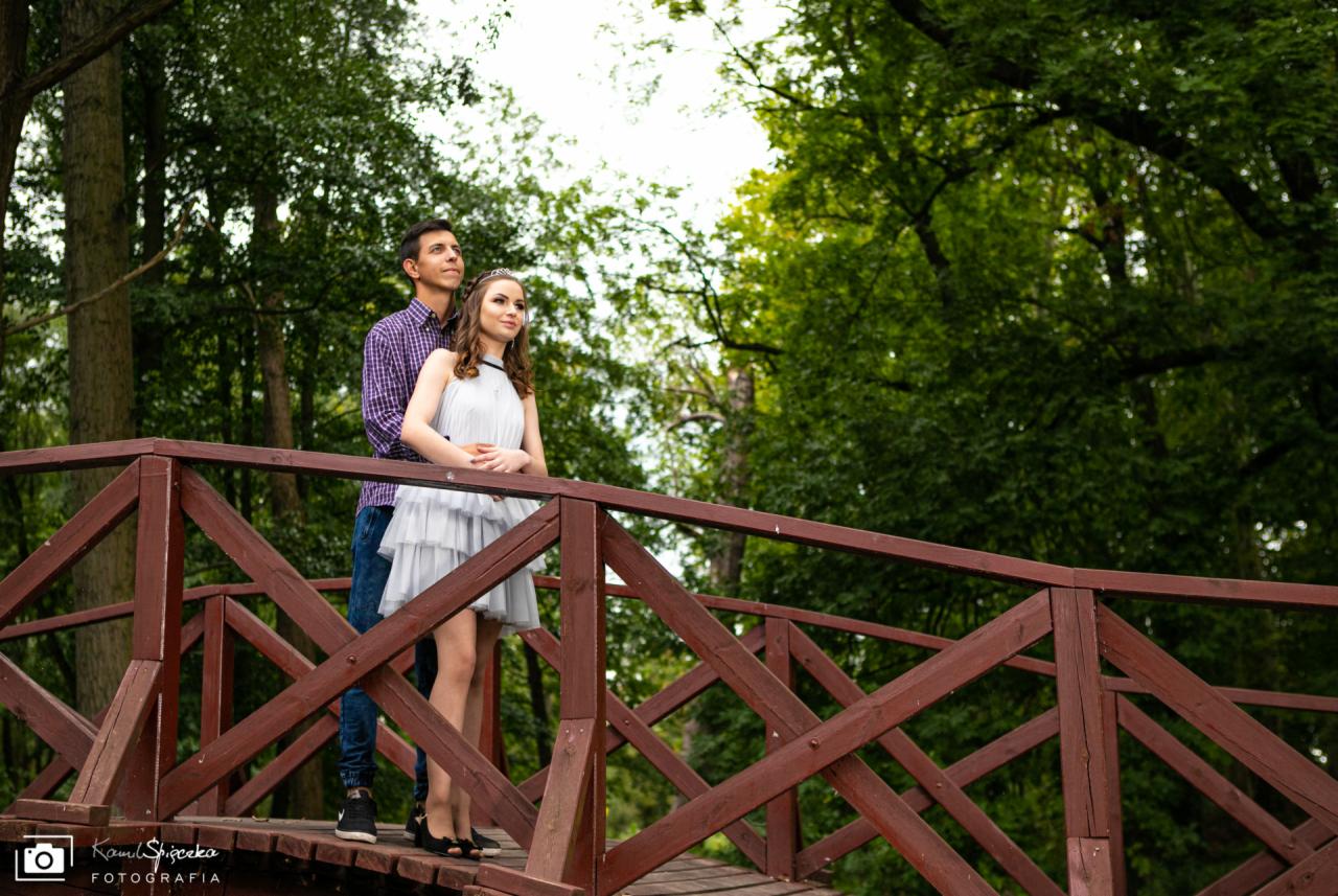 Para zakochanych stoi na drewnianym mostku w otoczeniu zieleni, spoglądając w dal; kobieta w jasnej sukience z falbanami, mężczyzna w koszuli w kratę.