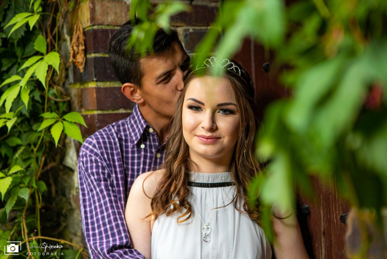 Młody mężczyzna w koszuli w kratę całuje w policzek uśmiechniętą kobietę z tiarą na głowie, na tle ceglanej ściany porośniętej bluszczem, częściowo zasłonięci liśćmi.