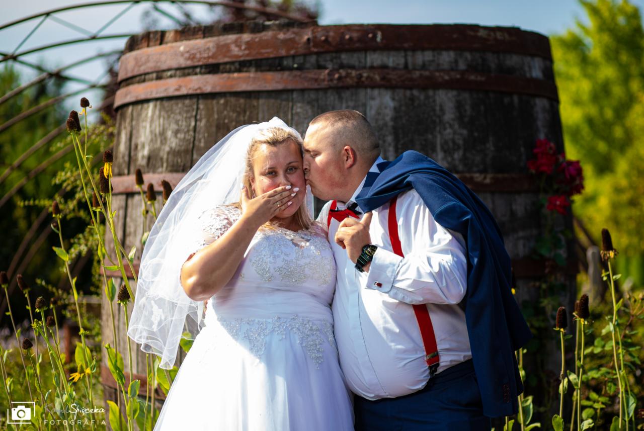 Panna młoda w białej sukni zakrywa usta dłonią, podczas gdy pan młody w białej koszuli z czerwonymi szelkami całuje ją w policzek na tle dużej drewnianej beczki i zieleni.