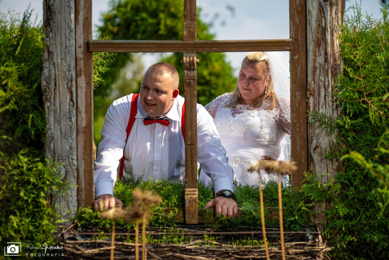 Para młoda pozuje za starą drewnianą ramą okienną porośniętą zielenią, on w białej koszuli, czerwonych szelkach i muszce, ona w białej sukni ślubnej z koronką i welonem.