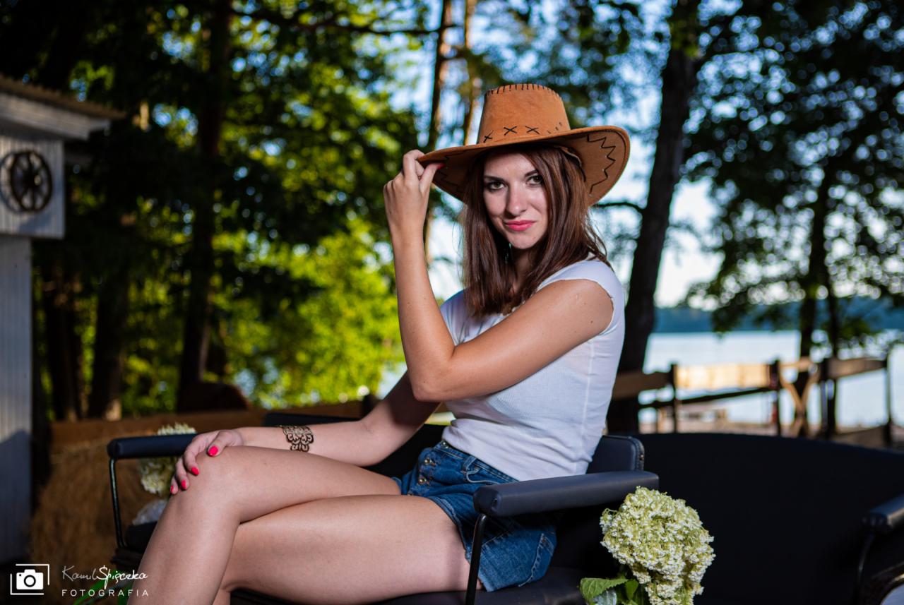 Kobieta w kapeluszu kowbojskim i dżinsowych szortach siedzi na czarnej ławce z kwiatami, pozując na tle drzew i jeziora.