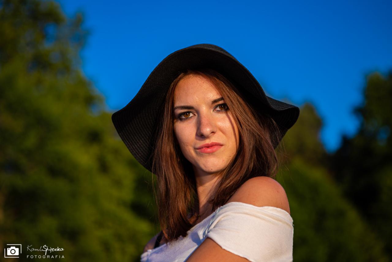 Portret młodej kobiety w czarnym kapeluszu z szerokim rondem, ubrana w białą bluzkę z odkrytymi ramionami, na tle zieleni i błękitnego nieba.