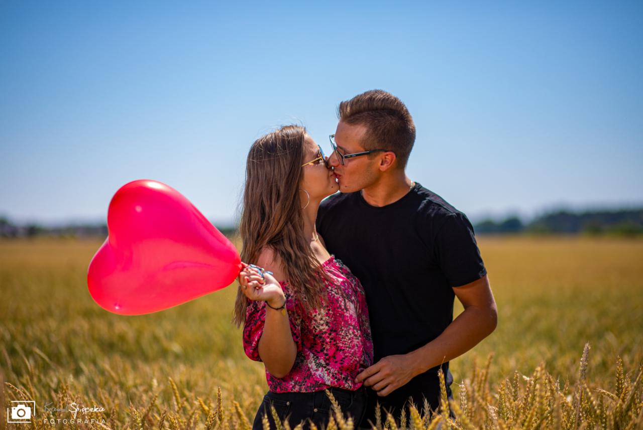 Zakochana para całuje się na tle pola zboża, trzymając czerwony balon w kształcie serca.