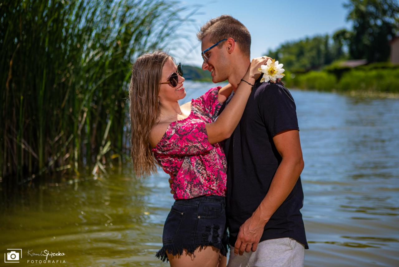 Uśmiechnięta para w okularach przeciwsłonecznych, obejmująca się na tle jeziora i trzcin, z kwiatem we włosach mężczyzny.