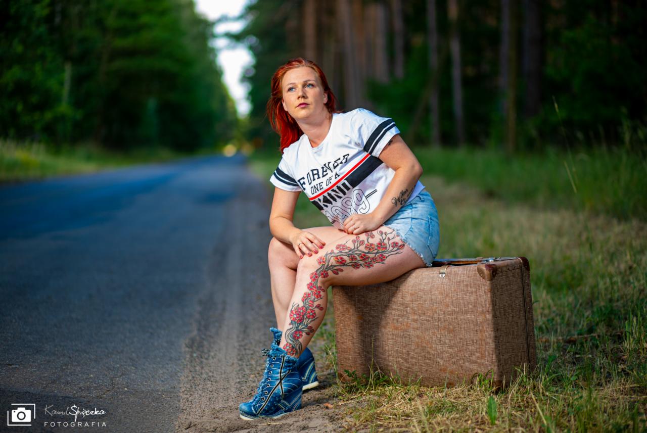 Kobieta z rudymi włosami i tatuażem kwiatowym na udzie siedzi na walizce przy drodze leśnej, ubrana w dżinsowe szorty i t-shirt.