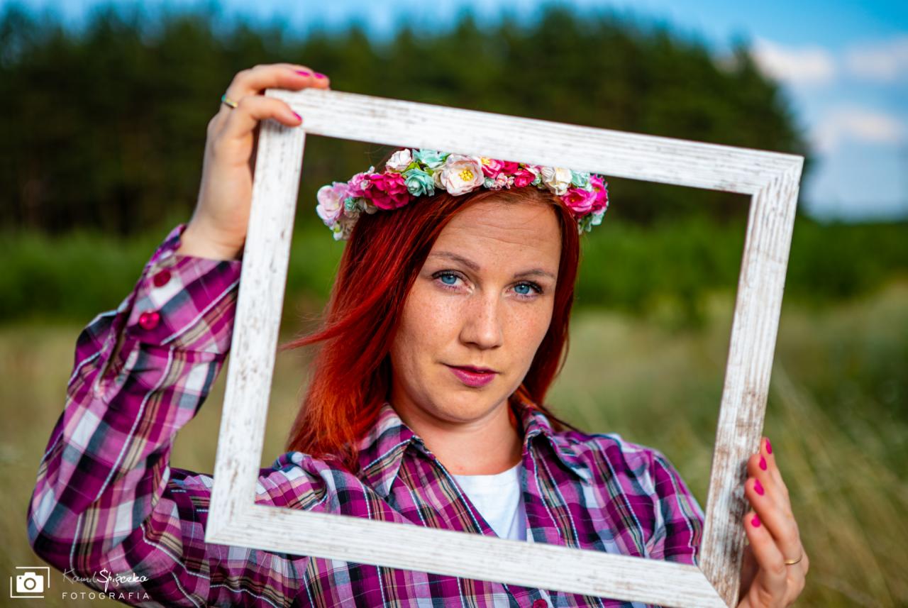 Portret kobiety z rudymi włosami, w kwiecistym wianku, trzymającej białą ramkę przed twarzą na tle zieleni.