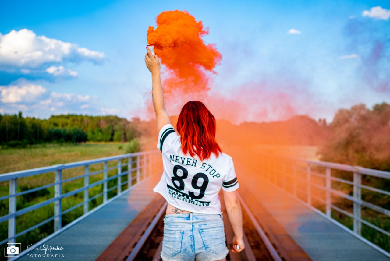 Kobieta z czerwonymi włosami, stojąca tyłem na moście w Międzyrzeczu, trzyma w górze odpaloną świecę dymną generującą pomarańczowy dym.