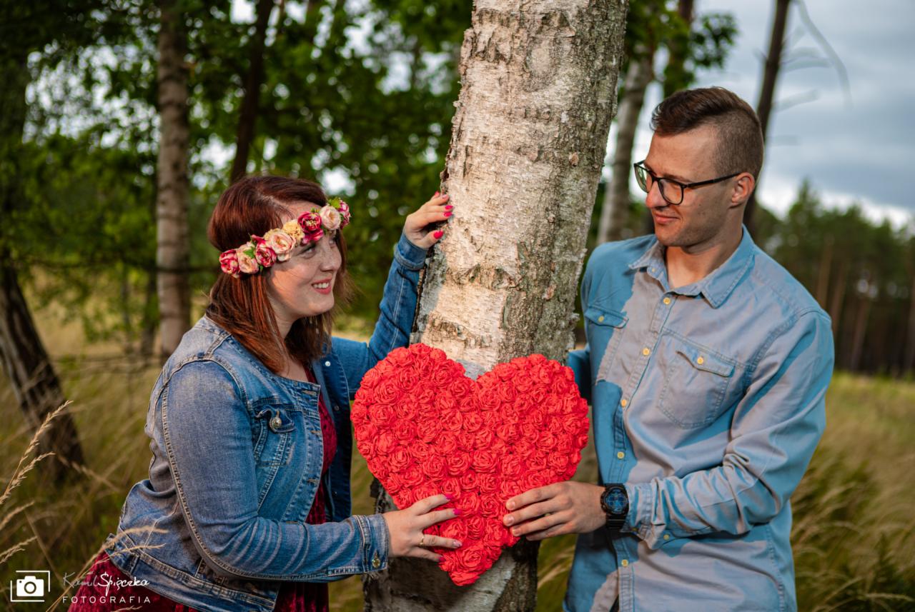 Uśmiechnięta para trzymająca czerwone serce z róż przy brzozie w plenerze, kobieta w wianku z kwiatów i dżinsowej kurtce, mężczyzna w okularach i błękitnej koszuli.