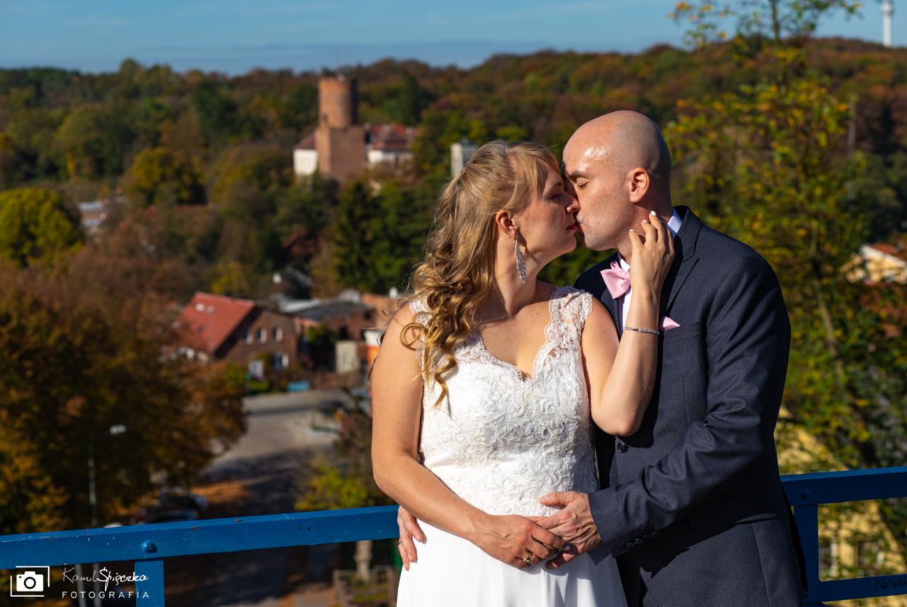 Pocałunek pary młodej na tle jesiennego krajobrazu z ceglaną wieżą w oddali, panna młoda w koronkowej sukni, pan młody w granatowym garniturze i różowej muszce.