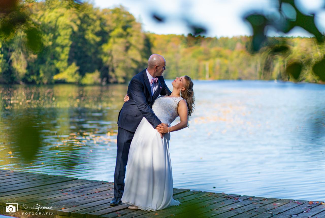 Para młoda w objęciach na drewnianym pomoście nad jeziorem w słoneczny dzień, mężczyzna w granatowym garniturze i różowej muszce, kobieta w białej sukni ślubnej z koronkową górą.