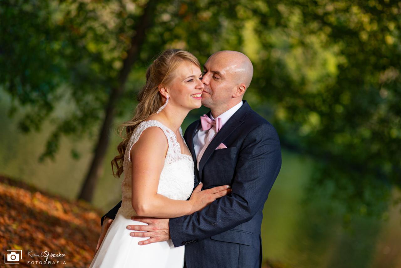 Uśmiechnięta panna młoda w koronkowej sukni, obejmowana przez pana młodego w granatowym garniturze z różową muchą, na tle jesiennych drzew i wody.