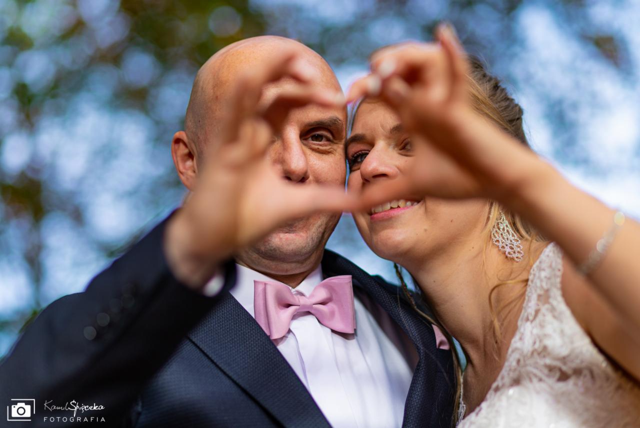 Uśmiechnięta para młoda tworzy dłońmi kształt serca na tle rozmytej zieleni, mężczyzna w granatowym garniturze z różową muchą, kobieta w białej koronkowej sukni i długich kolczykach.