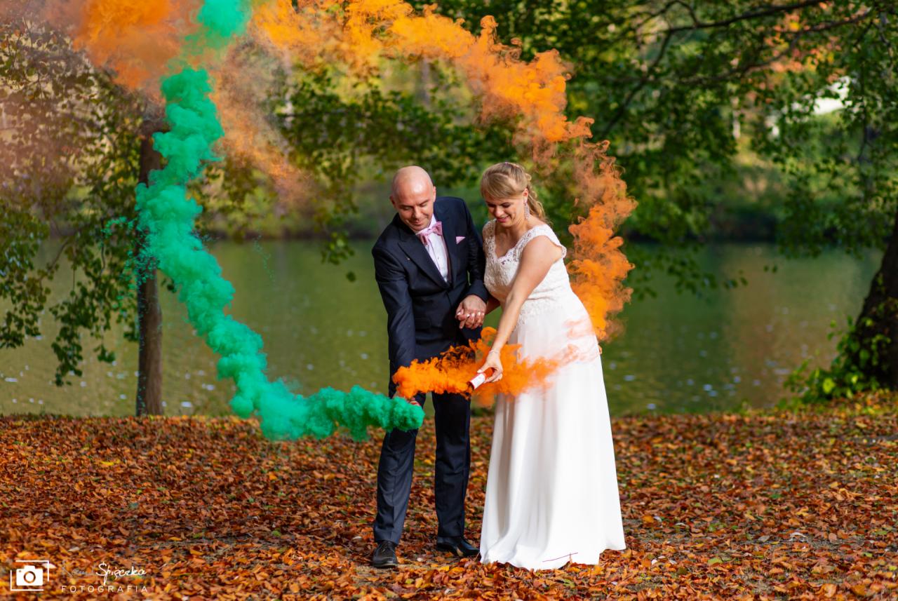 Para młoda w strojach ślubnych trzymająca dymne świece w kolorach zielonym i pomarańczowym na tle jesiennego krajobrazu z jeziorem.