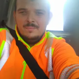 Mężczyzna w pomarańczowej kurtce roboczej z elementami odblaskowymi, zapięty pasem bezpieczeństwa w samochodzie, z widocznym fragmentem okna.