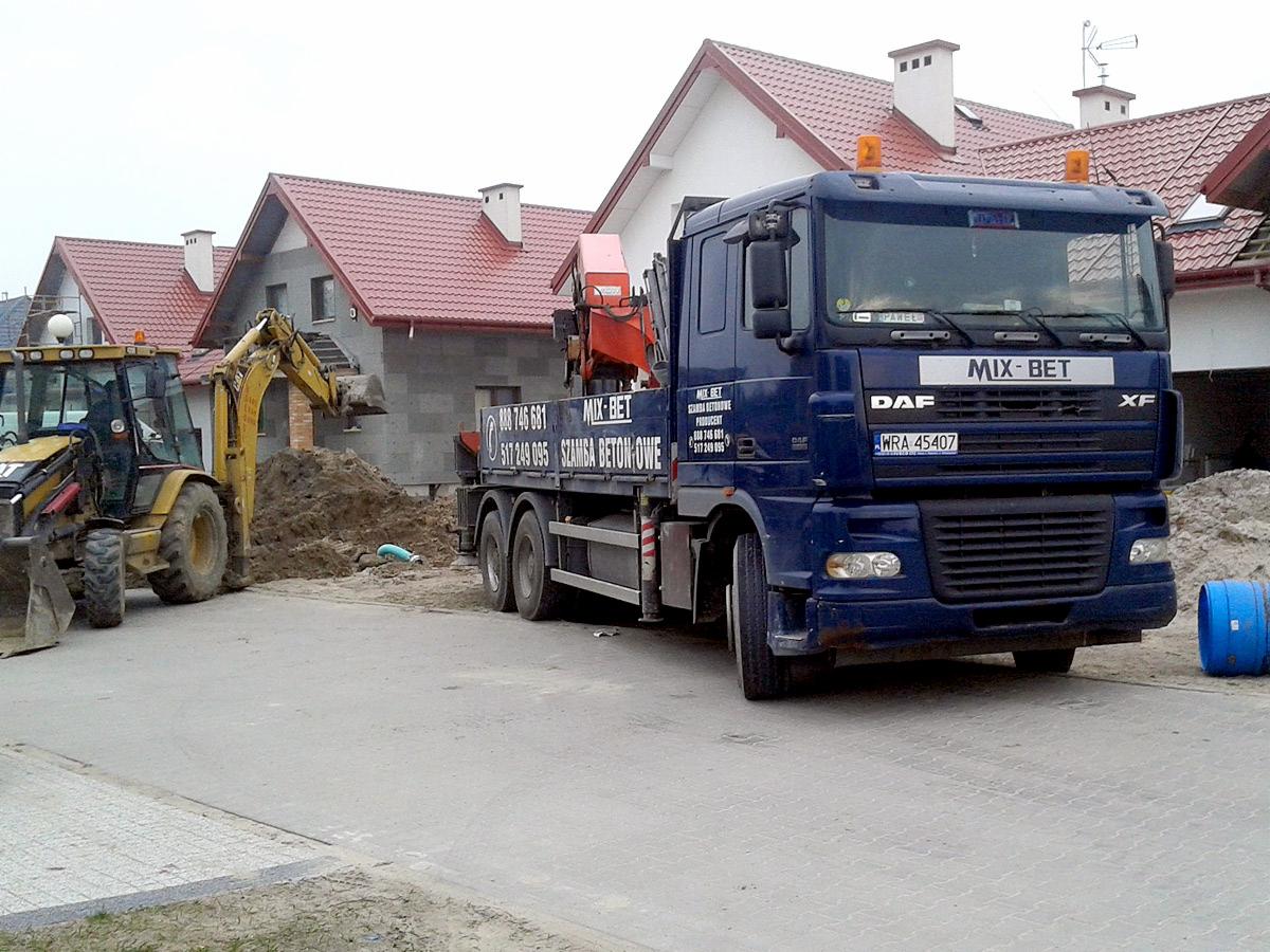 Żółta koparka i niebieska ciężarówka z dźwigiem na tle budynków w trakcie budowy, widoczny wykop i materiały budowlane.