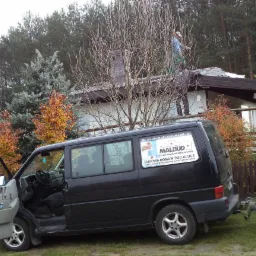 Dwóch pracowników na dachu jednopiętrowego domu podczas prac, widoczny czarny bus z przyczepką i reklamą firmy budowlanej na boku, otwarte drzwi kierowcy, jesienne drzewa w tle.