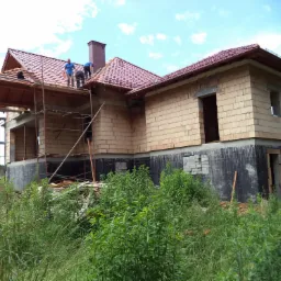 Budowa domu jednorodzinnego w trakcie realizacji: wzniesione mury z pustaków, częściowo położona dachówka ceramiczna, widoczne rusztowania i pracujący dekarze, wysoka trawa na pierwszym planie.