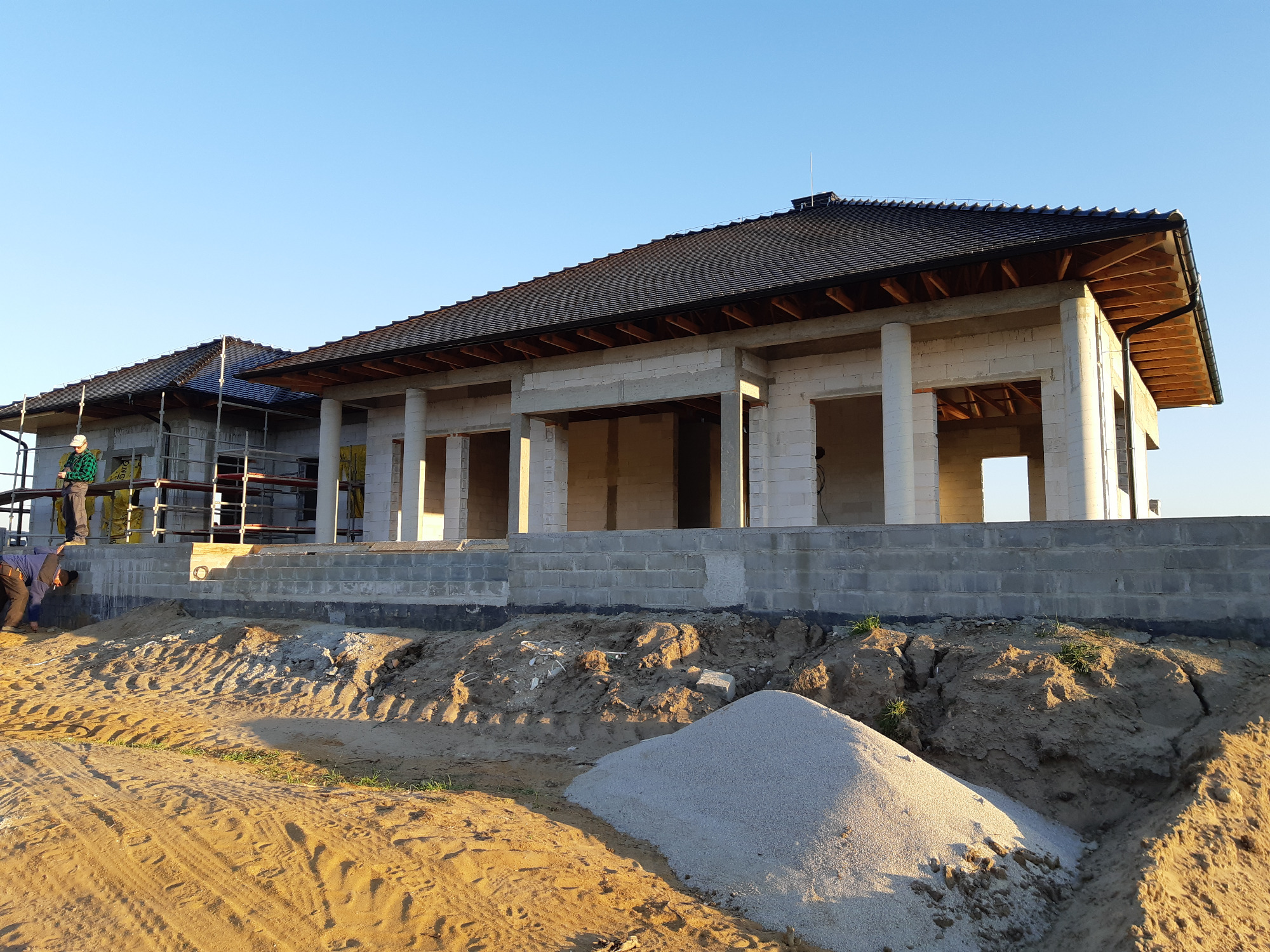 Surowy stan domu z pustaków z ciemną dachówką, widoczny stos piasku i pracownicy na rusztowaniu w pogodny dzień.