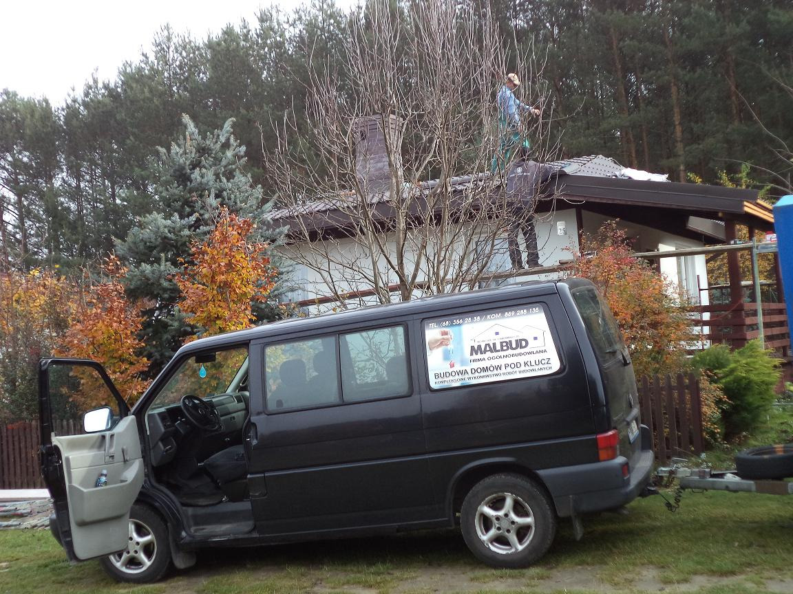 Dwóch pracowników na dachu jednopiętrowego domu podczas prac, widoczny czarny bus z przyczepką i reklamą firmy budowlanej na boku, otwarte drzwi kierowcy, jesienne drzewa w tle.