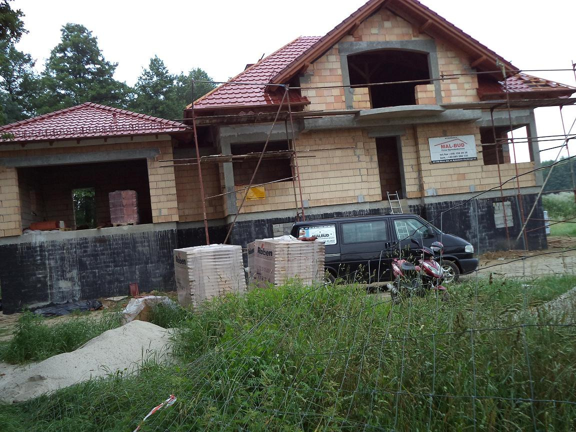 Niedokończony budynek z czerwonej cegły z częściowo ułożonym czerwonym dachem, widoczne rusztowania, zaparkowany czarny van z logo firmy budowlanej i skuter, otoczony zielenią.