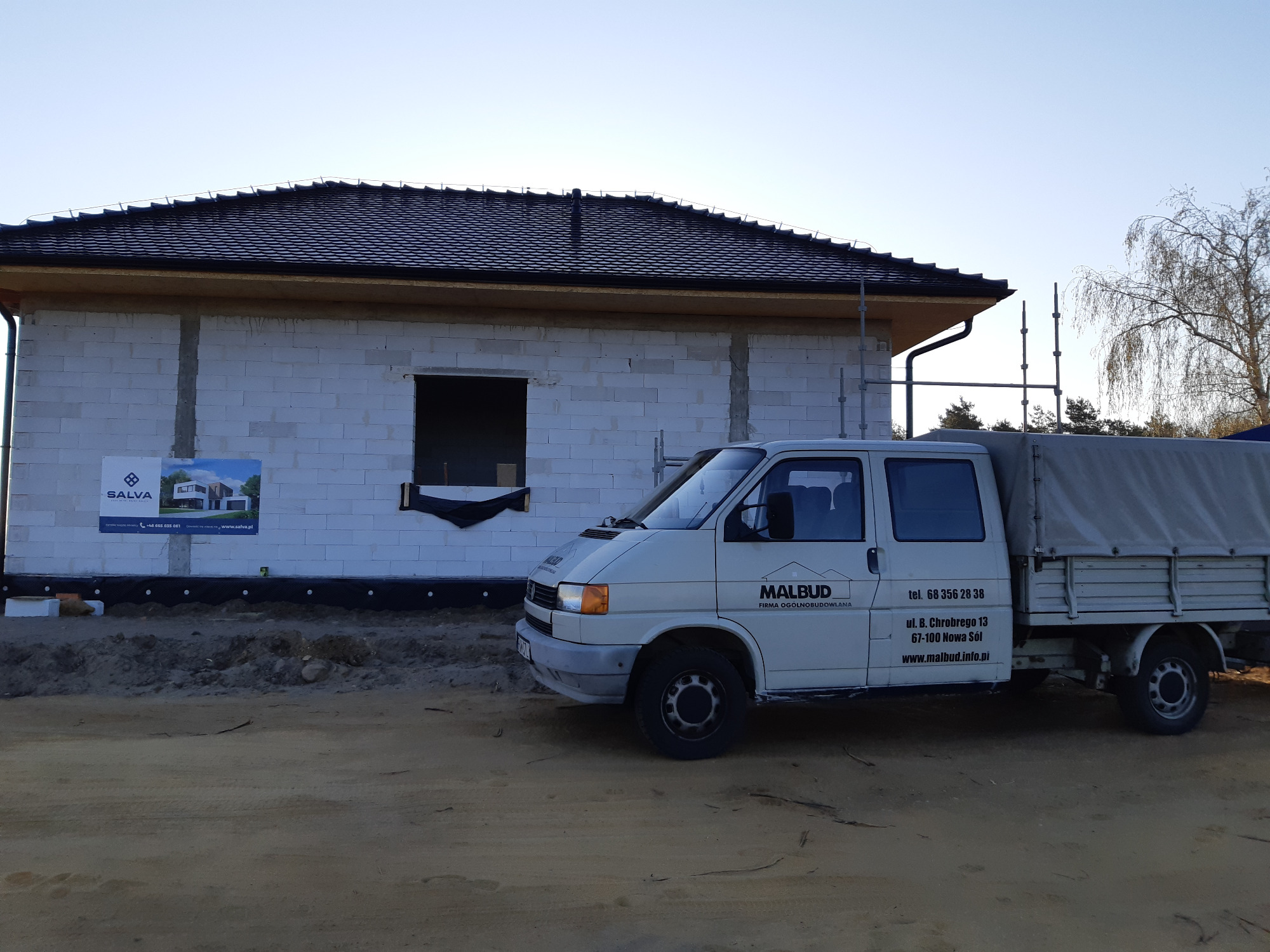 Surowy stan domu z białego bloczka, z czarnym dachem i widocznym oknem, na tle którego stoi biały samochód dostawczy z logo firmy budowlanej Malbud.