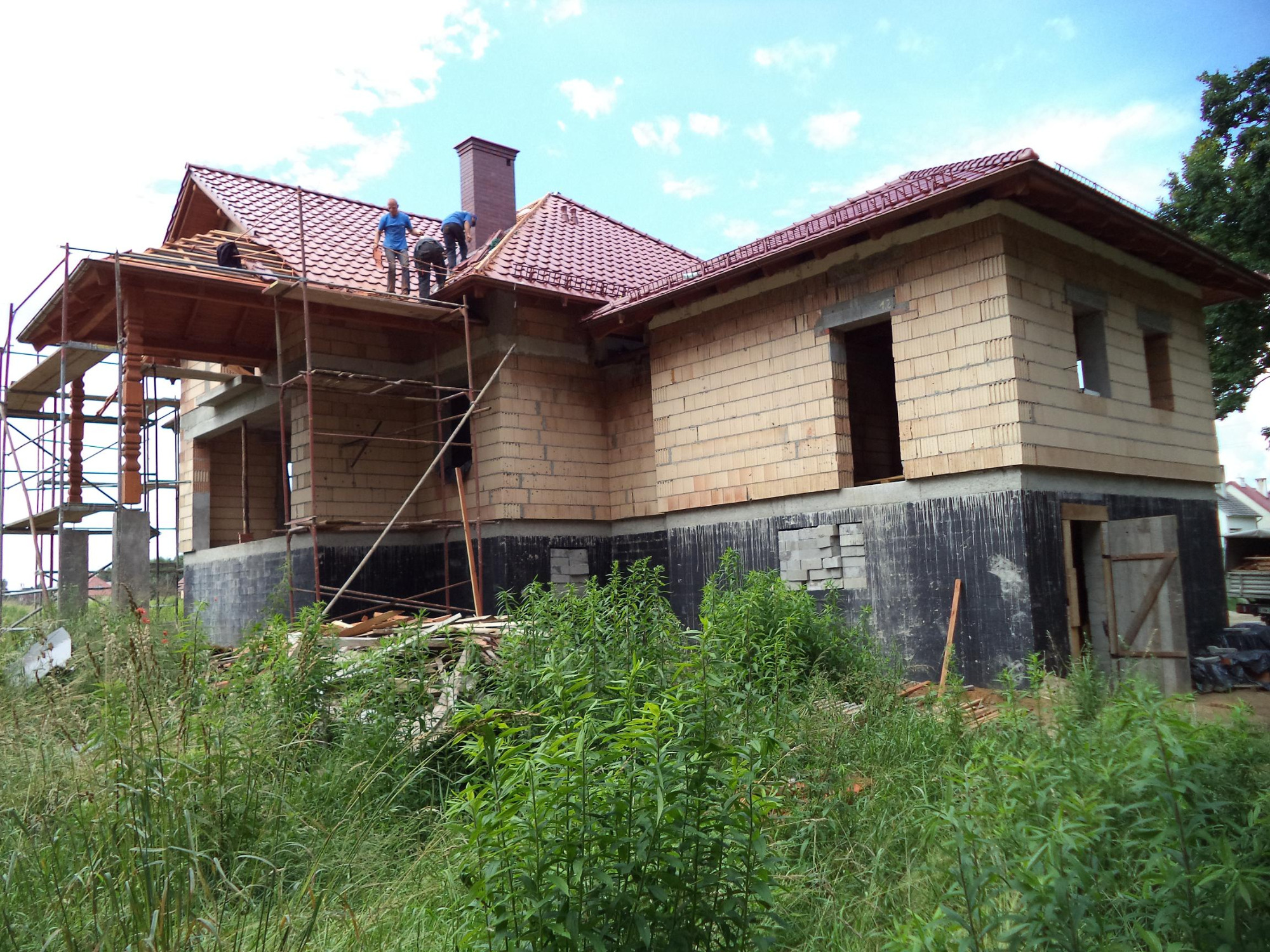 Budowa domu jednorodzinnego w trakcie realizacji: wzniesione mury z pustaków, częściowo położona dachówka ceramiczna, widoczne rusztowania i pracujący dekarze, wysoka trawa na pierwszym planie.