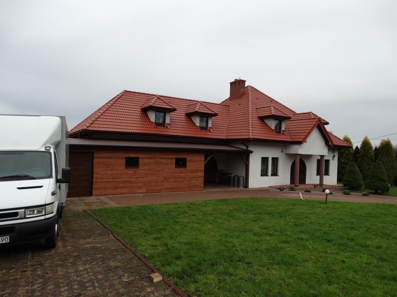Dostawczy samochód na podjeździe przed domem z czerwoną dachówką i drewnianą elewacją garażu, widoczne kosze na śmieci.