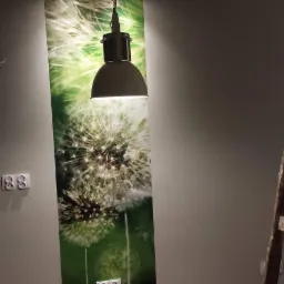 Ściana z fototapetą przedstawiającą dmuchawce, oświetlona wiszącą lampą. Widoczne gniazdka elektryczne, wałek do malowania i drabina.