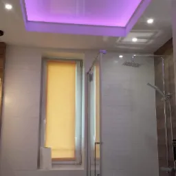 Nowoczesna łazienka z podwieszanym sufitem podświetlonym na fioletowo, kabiną prysznicową ze szklanymi drzwiami i roletą w oknie.