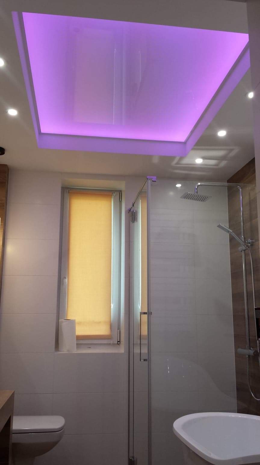 Nowoczesna łazienka z podwieszanym sufitem podświetlonym na fioletowo, kabiną prysznicową ze szklanymi drzwiami i roletą w oknie.