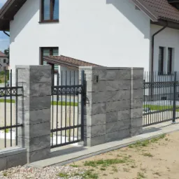 Nowoczesne ogrodzenie z paneli betonowych w kolorze szarości z czarną metalową bramką i przęsłami, na tle białego domu z brązowym dachem, widok z zewnątrz posesji.
