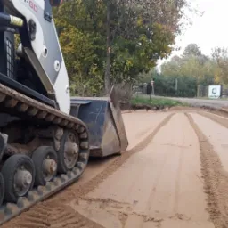 Kompaktowa ładowarka gąsienicowa wyrównuje teren piaskiem, widoczne ślady gąsienic i lemiesz, w tle drzewa i tablica informacyjna.