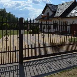 Ciemna, metalowa brama wjazdowa z ozdobnymi elementami, prowadząca na wybrukowany podjazd przed domem z białymi ścianami i ciemnym dachem.