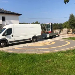 Biały bus z przyczepą przewożącą minikoparkę stoi na okrągłym podjeździe z kostki brukowej w żółto-szare wzory, przed budynkiem i stertą materiałów budowlanych.