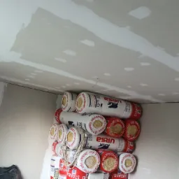 Stos rolek wełny mineralnej URSA Standard, czekających na montaż w pomieszczeniu z częściowo wykończonymi ścianami z płyt kartonowo-gipsowych i surowym, niepomalowanym sufitem.