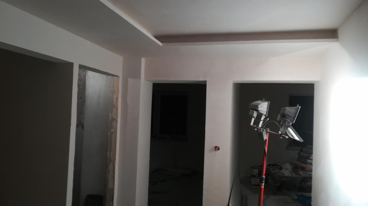 Wnętrze w trakcie malowania: świeżo pomalowane ściany na biało, widoczne otwory drzwiowe, lampa budowlana na statywie, worki z materiałami budowlanymi w tle.