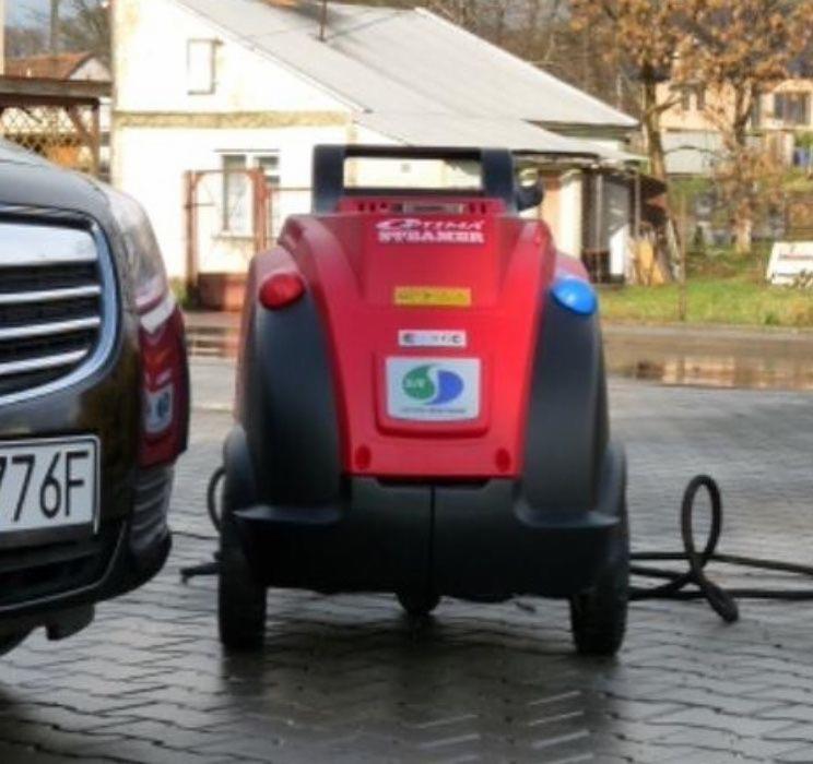 Czerwono-czarna parownica mobilna Optima Steamer na zewnątrz, obok zaparkowanego samochodu, z widocznym przewodem na mokrej nawierzchni.