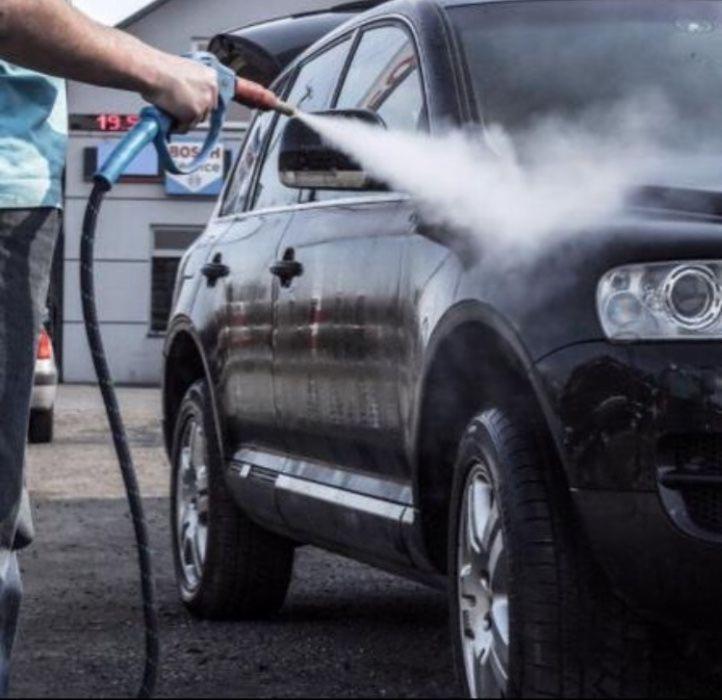 Czarny SUV myty pod ciśnieniem przez osobę trzymającą lancę z dyszą, widoczny budynek myjni w tle.