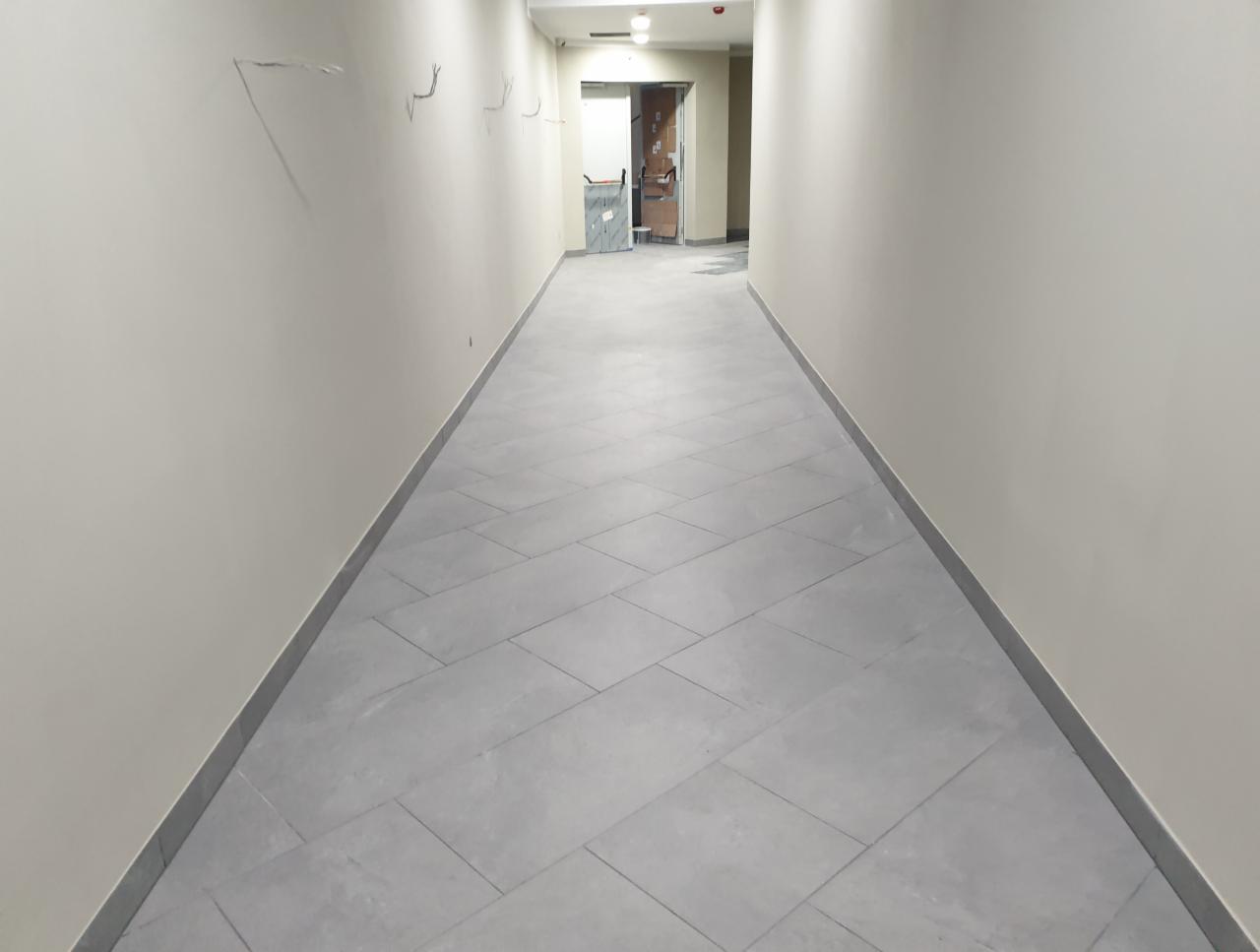Długi korytarz z ułożoną szarą płytką podłogową w jodełkę, ściany w kolorze jasnego beżu z widocznymi śladami po instalacjach elektrycznych, w oddali widoczny wózek transportowy i otwarte drzwi.