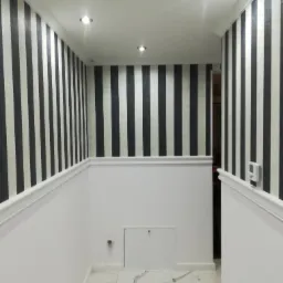 Wnętrze korytarza z tapetą w czarno-białe pionowe pasy, białą lamperią i marmurową podłogą. Widoczne oświetlenie punktowe w suficie i białe listwy przypodłogowe.