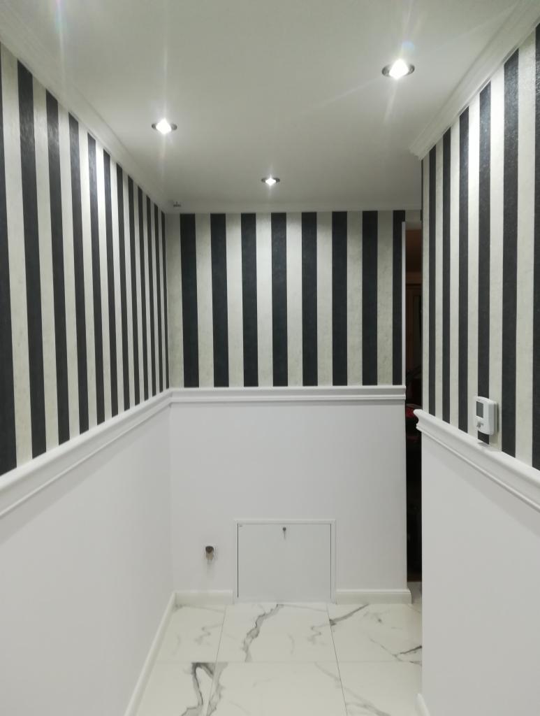 Wnętrze korytarza z tapetą w czarno-białe pionowe pasy, białą lamperią i marmurową podłogą. Widoczne oświetlenie punktowe w suficie i białe listwy przypodłogowe.