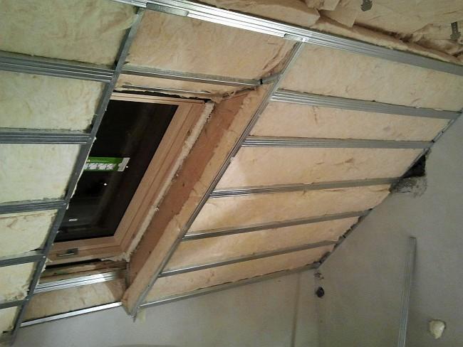 Skośny sufit poddasza w trakcie izolacji wełną mineralną, widoczne metalowe profile i okno dachowe.