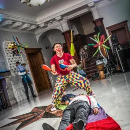 Klaun w kolorowych spodniach żongluje maczugami nad leżącym mężczyzną na kolorowej chuście, w tle dekoracje balonowe i publiczność.