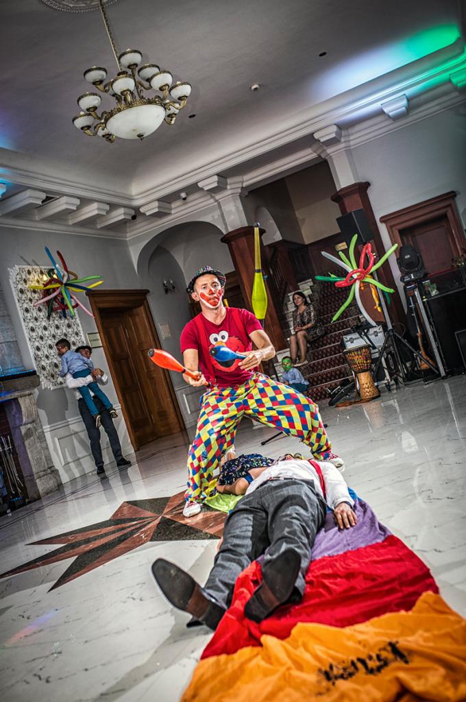 Klaun w kolorowych spodniach żongluje maczugami nad leżącym mężczyzną na kolorowej chuście, w tle dekoracje balonowe i publiczność.