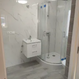 Nowoczesna łazienka po remoncie: kabina prysznicowa z zaokrąglonym szkłem, biała umywalka z szafką i marmurowe płytki ścienne z szarymi żyłami.