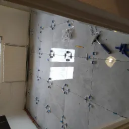 Układanie jasnoszarych płytek podłogowych z użyciem systemu poziomowania, widoczne narzędzia: młotek, ołówek i szczypce do klinów, w tle okno z zielenią.